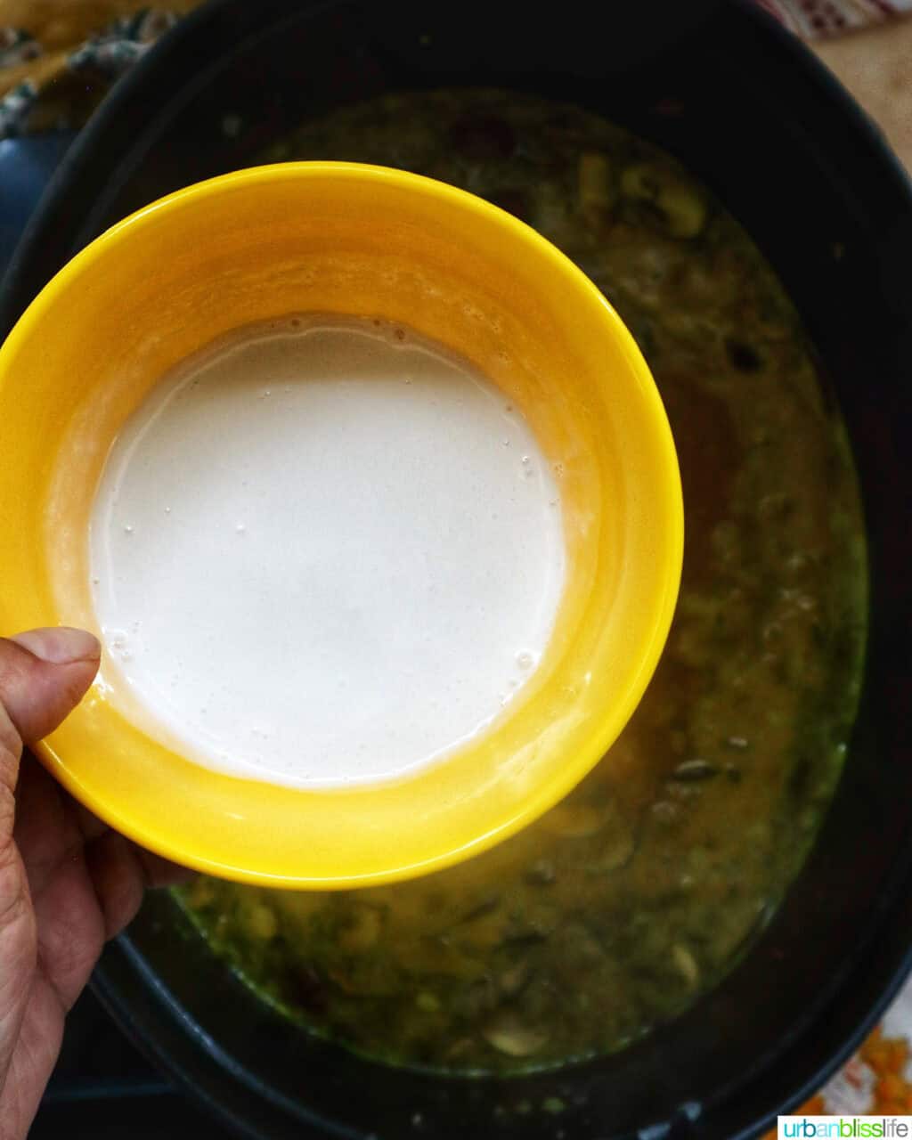 hand holding a yellow bowl of whipping cream over a dutch oven of soup.