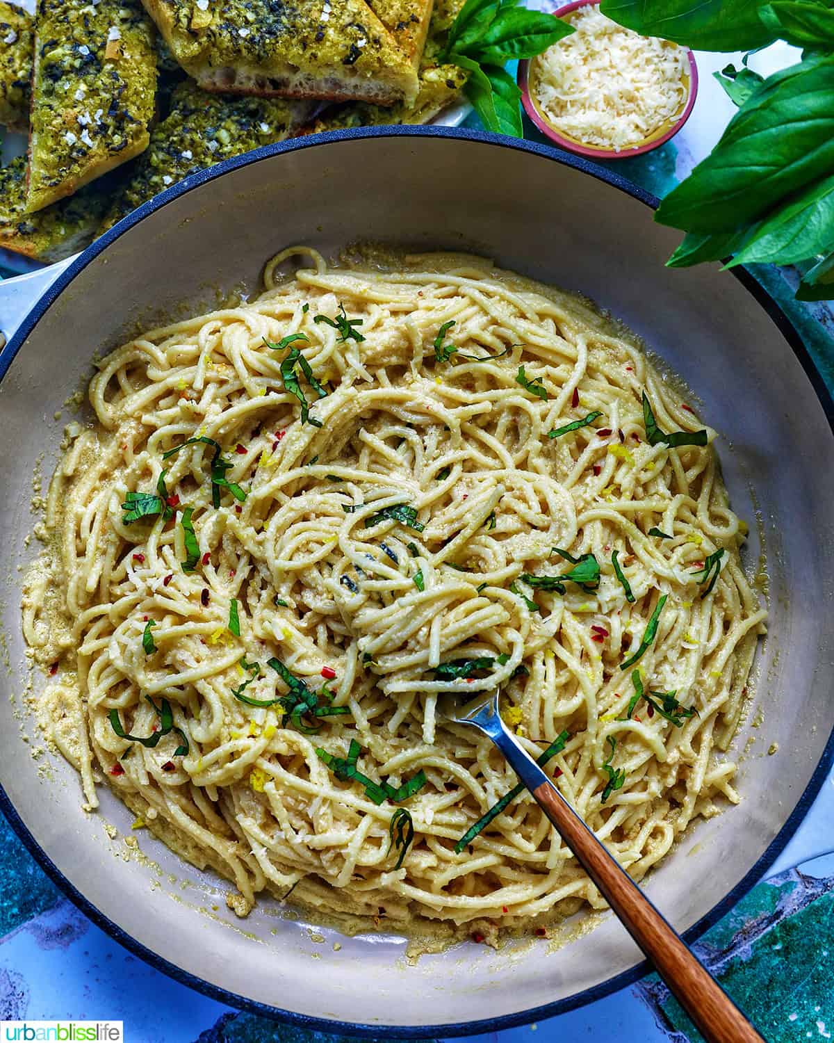 saucepan of tahini pasta with plate of pesto bread and side bowls of pine nuts, cheese, and parsley.