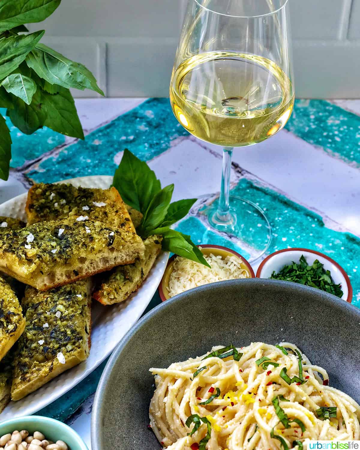 bowl of tahini pasta with plate of pesto bread and side bowls of pine nuts, cheese, and parsley with glass of white wine.