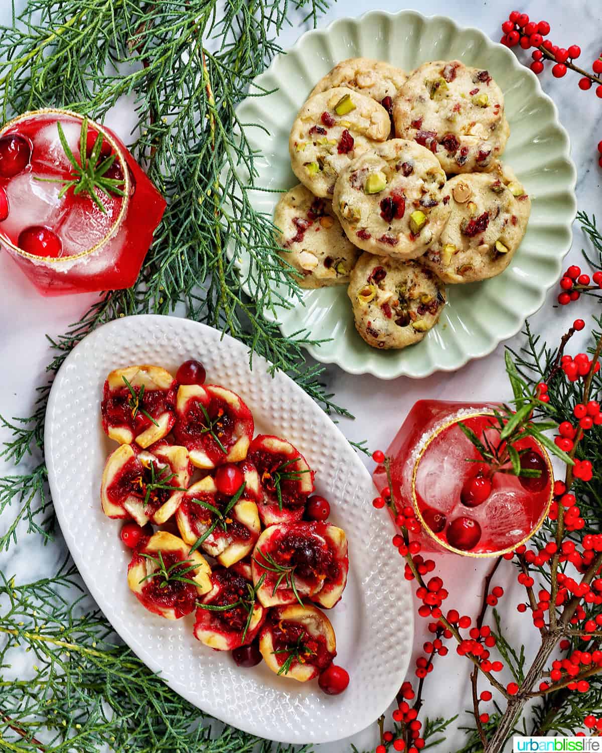 cranberry brie tartlet appetizers on a plate next to plate of pistachio cranberry shortbread cookies and glasses of cranberry mocktails with holiday decor.