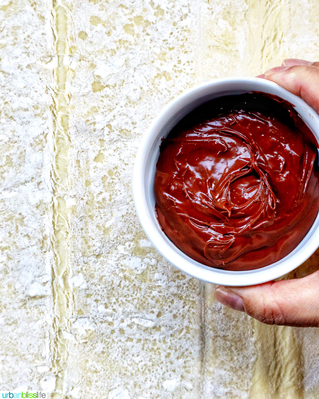 hand holding a bowl of chocolate over puff pastry dough.