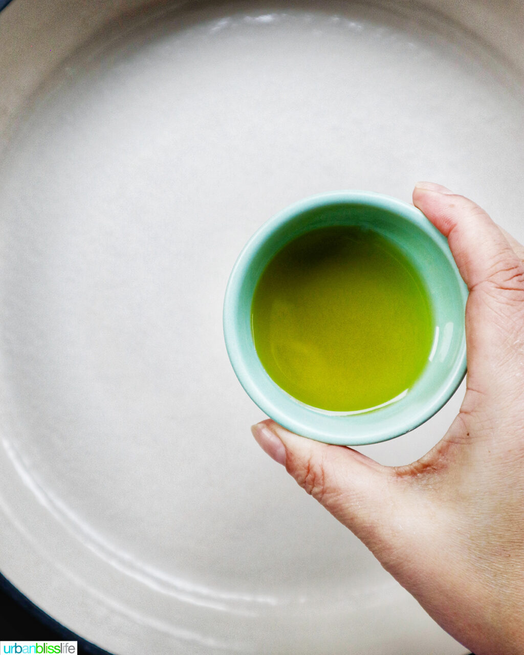 hand holding bowl of olive oil over pan.