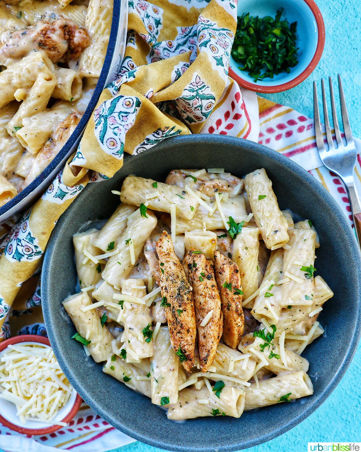Bowl of garlic chicken parmesan pasta with side bowl of parsley next to pan of pasta.