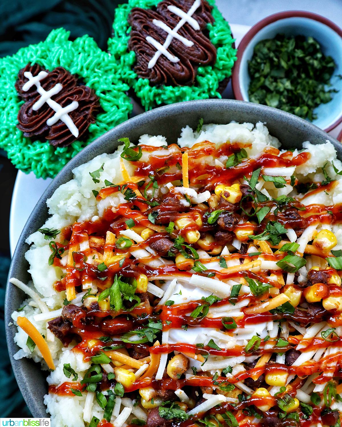 loaded potato bowl with mashed potatoes, beans, corn, cheese, and herbs next to football cupcakes.