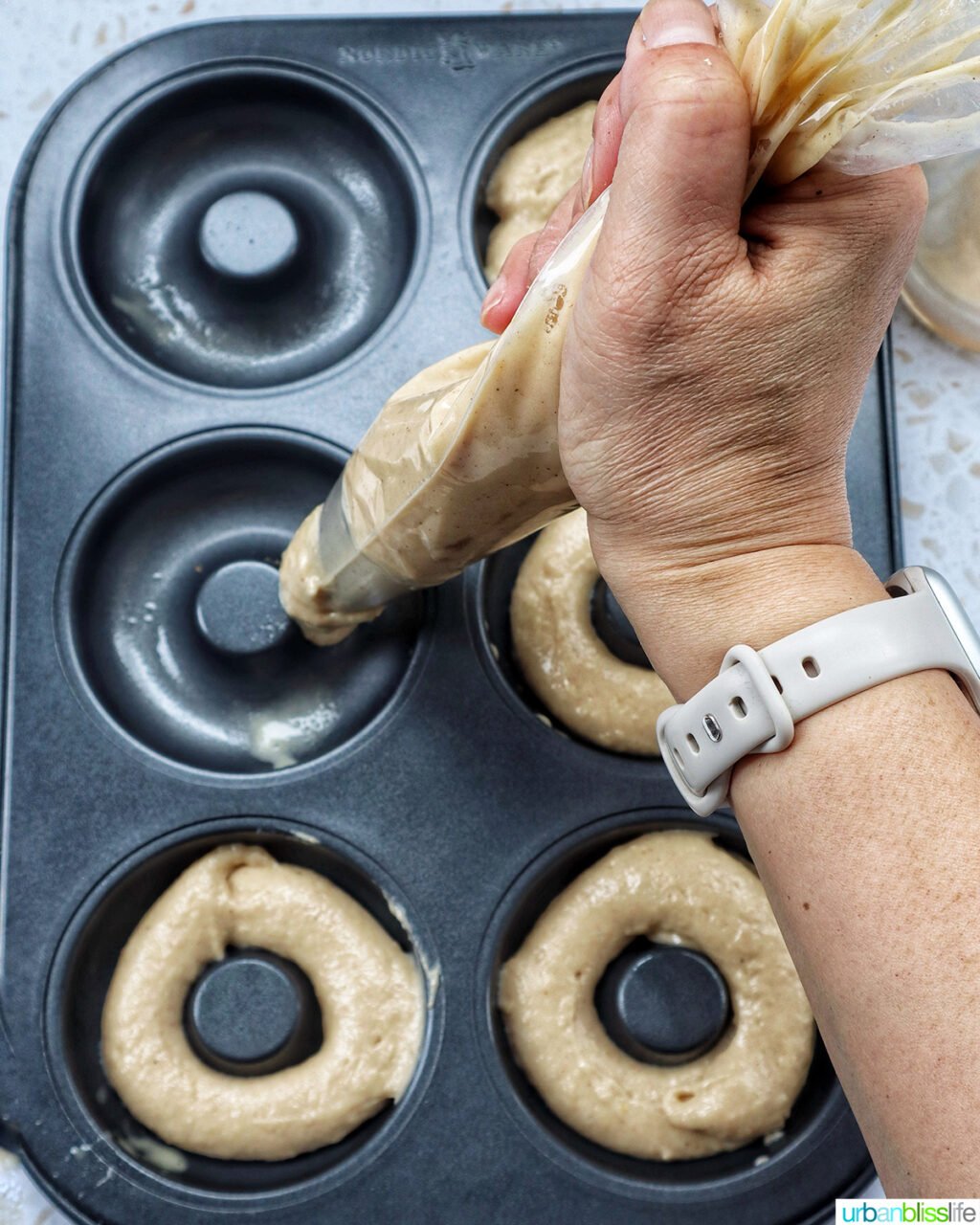 hand holding a piping bag piping churro donut batter into donut pan.