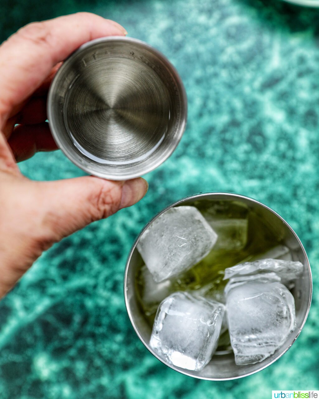 hand holding a cocktail jigger over a cocktail shaker filled with ice.