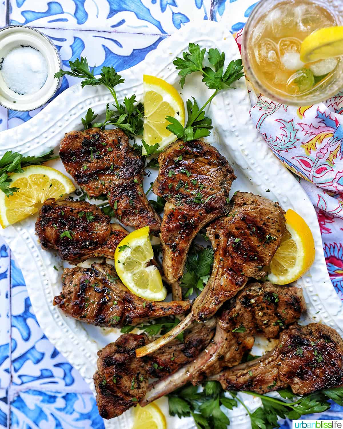 Greek lamb chops on a white plate with herbs and lemon slices and bourbon lemonade.