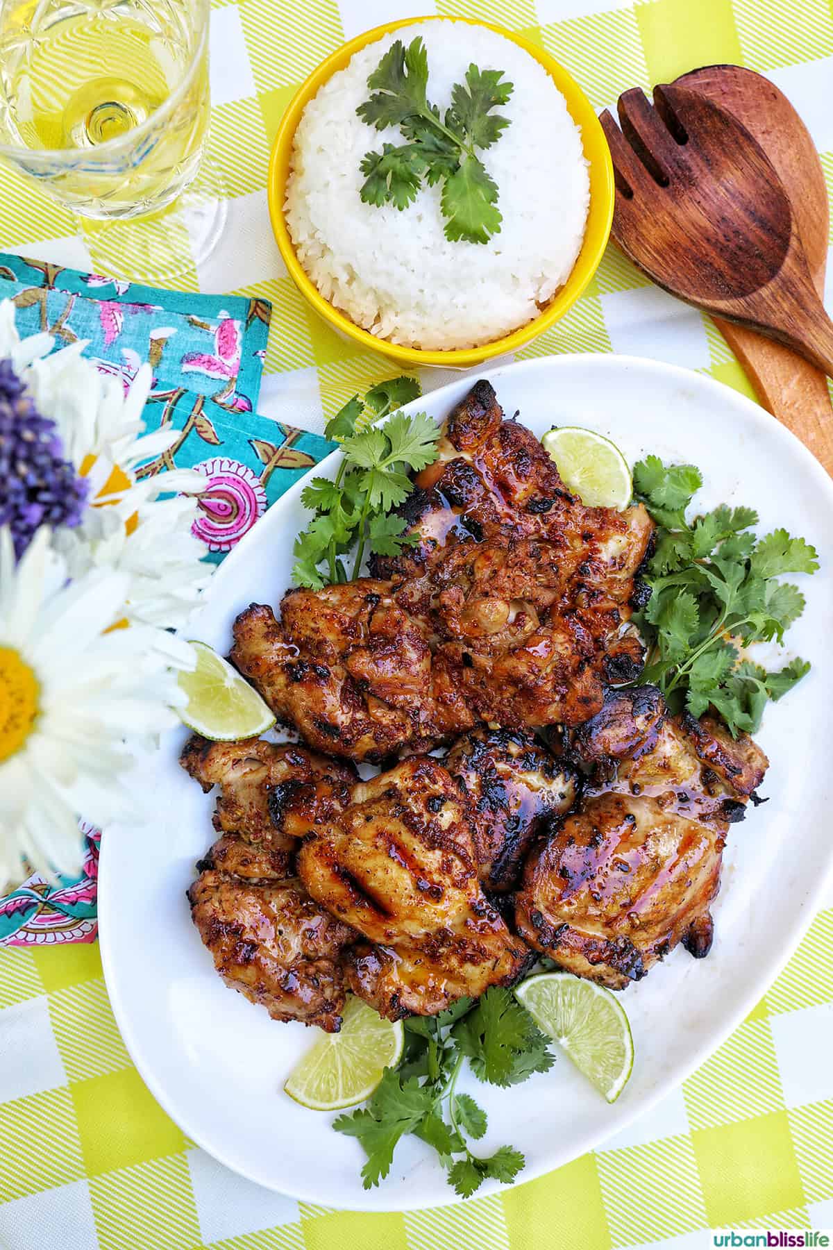 Filipino grilled chicken inasal on a platter near a vase of daisies and bowl of rice.